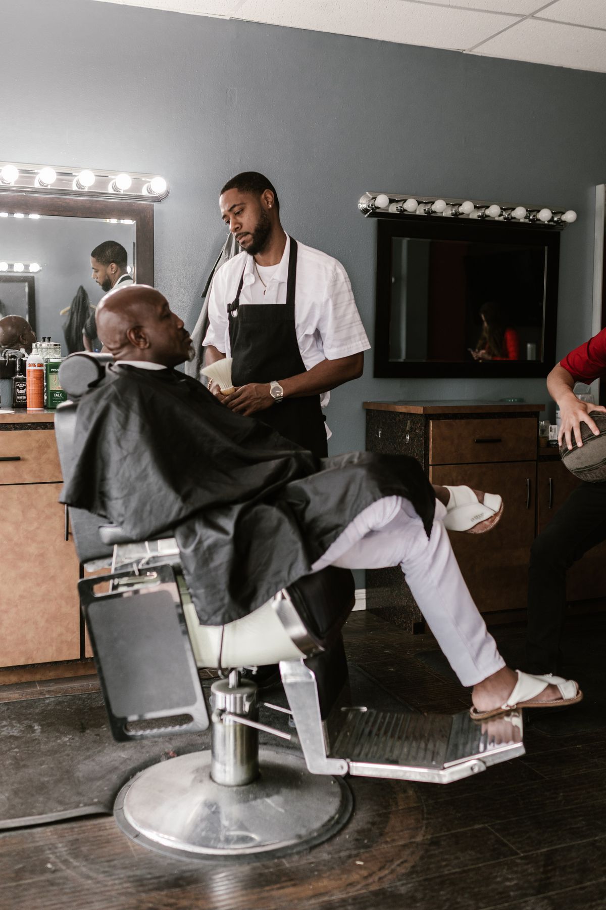 A barber working with a seated customer in a neighborhood barbershop, with mirrors and tools at the workstation.